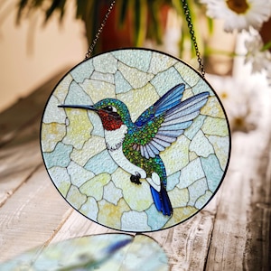 Könnte beinhalten: Ein rundes Buntglasstück mit einem Kolibri mit ausgebreiteten Flügeln. Der Vogel hat einen roten Hals, einen grünen Körper und blaue Flügel. Der Hintergrund ist ein Mosaik aus hellblauen, weißen und gelben Formen, umrahmt von einem schwarzen Metallrand und einer Kette.