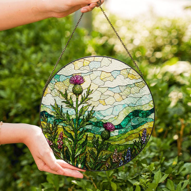 A hand holds a round stained glass piece featuring a vibrant landscape scene with a thistle and other flowers.