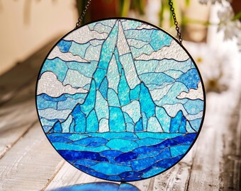 Può includere: Una finestra in vetro colorato che rappresenta una catena montuosa blu con un cielo bianco e nuvole. La catena montuosa è circondata da un oceano blu. La finestra è rotonda e ha una cornice metallica.