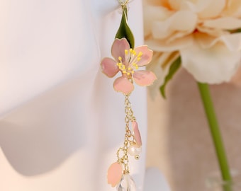 Op de afbeelding: Een lange bungelende oorbel met een roze emaille kersenbloesem en een groen blaadje, hangend aan een goudkleurige ketting. De ketting bevat extra kleine roze bloemblaadjes, een witte parel en een heldere kristallen druppel als hanger.