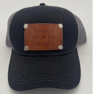 May include: Black and gray trucker hat with a brown leather patch that reads "Horses Keep Me Stable".  The hat comes with one patch.  Other patch options include "Y'all Need Groundwork", "I Will Not Hold My Horses", "Ride More Worry Less", "Horse Show Squad", "If You Can Read This Put Me Back On The Horse", and "Barn Hair Don't Care".