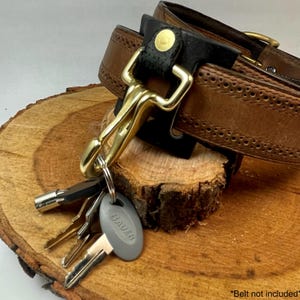 Könnte beinhalten: Ein Schlüsselhaken aus Messing, der an einem braunen Ledergürtel mit einem schwarzen Lederflicken befestigt ist. Der Haken hält einen Satz Schlüssel mit einem grauen Schlüsselanhänger, auf dem "BAUER" steht. *Gürtel nicht im Lieferumfang enthalten*