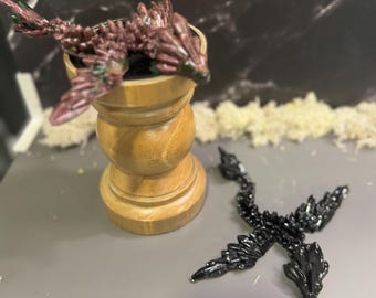 May include: Two miniature dragon figurines, one is black and the other is a reddish-purple colour with green accents. The purple dragon is perched on a wooden pedestal.