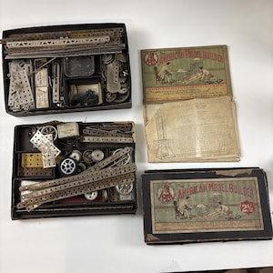 May include: Vintage metal construction sets, including two boxes filled with metal strips, gears, and other components. The boxes are accompanied by instruction manuals labeled "The American Model Builder."