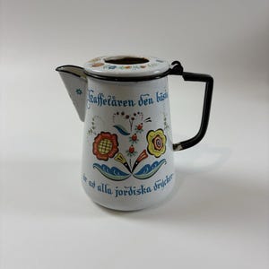 May include: A white ceramic mug with a black handle and the text "But First, Coffee" in a bold, black font. The mug is set against a blurred background, suggesting a cozy setting.