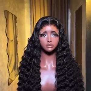 May include: A black curly wig styled on a mannequin head. The wig features long, voluminous curls and a natural-looking hairline. The mannequin has makeup applied, including eyelashes and eyeshadow.