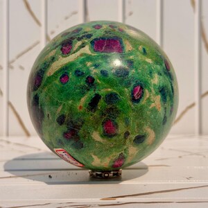 May include: A polished sphere of green zoisite mineral featuring embedded deep magenta ruby crystals and dark black hornblende inclusions. The surface displays a smooth, rounded texture with a small white sticker containing text.