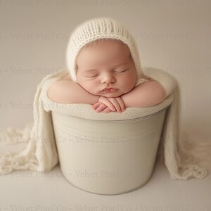 May include: A newborn baby wearing a white knitted hat sleeps peacefully in a white bucket, wrapped in a white blanket.