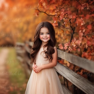 May include: A young girl with long brown hair smiles, wearing a light peach-colored dress, leaning against a wooden fence. The background features autumn foliage in shades of orange and red, creating a warm, seasonal scene.