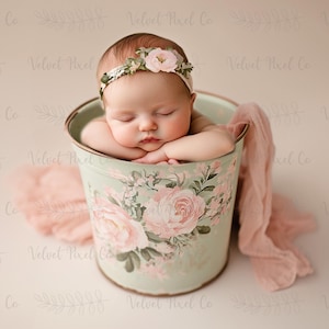 Op de afbeelding: Een pasgeboren baby slaapt in een groene, met bloemen geprinte emmer. De baby draagt een wit hoofdband met roze bloemen.