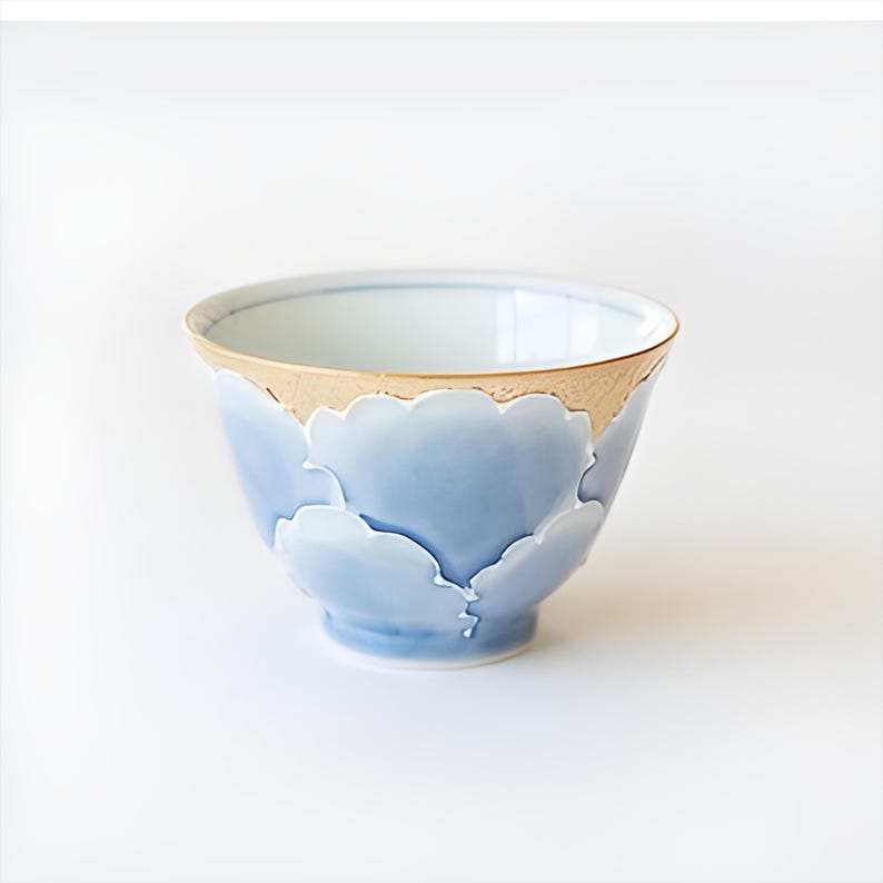 May include: A small, light blue ceramic bowl with a gold rim and a design of stylized clouds. The bowl is perfect for serving tea or other small dishes.