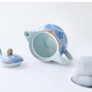 May include: A blue and white ceramic teapot with a floral design. The teapot has a lid and a strainer. The teapot is sitting on a white surface.
