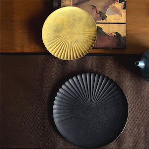 May include: Two round decorative plates, one black and one gold, with a fan-shaped design. The plates are on a brown surface.