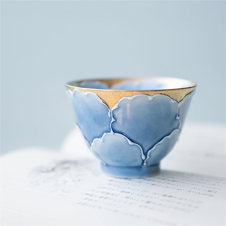 May include: A small, light blue ceramic teacup with a gold rim and a decorative pattern of stylized clouds. The cup is sitting on a white surface.