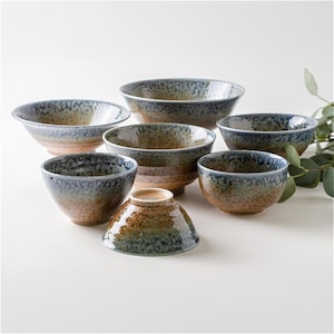 May include: A set of seven ceramic bowls with a speckled blue, brown, and tan glaze. The bowls vary in size and shape, with some having a rounded form and others a more angular design. The bowls are arranged on a white surface.