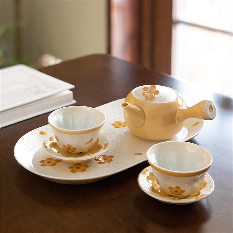 May include: A yellow ceramic teapot with a floral design on the lid, two matching teacups and saucers, and a white oval tray with a floral pattern.