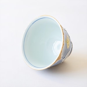 May include: A white ceramic teacup with a blue rim and a gold rim. The cup has a light blue glaze with a white design on the outside. The cup is sitting on a white surface.