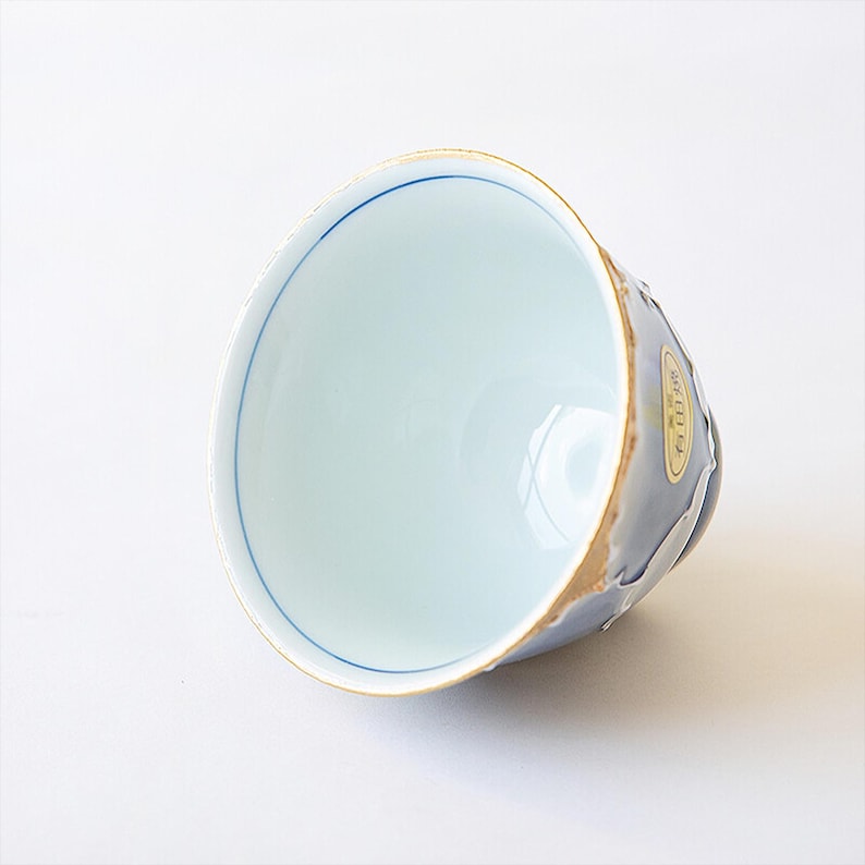May include: A white ceramic teacup with a blue rim and a gold rim. The cup has a light blue glaze with a white design on the outside. The cup is sitting on a white surface.