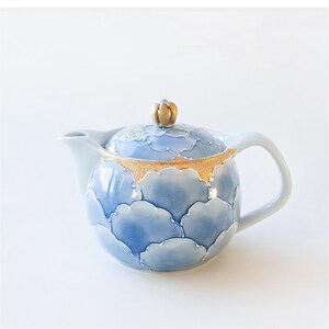 May include: A blue and gold ceramic teapot with a floral design. The teapot has a round body and a lid with a knob on top. The teapot is decorated with a pattern of blue flowers and gold accents.