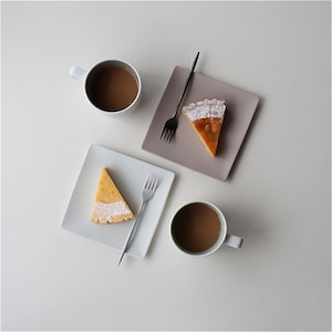 May include: Overhead shot of a breakfast or dessert setting. Two white mugs filled with coffee, two square plates with slices of pie, and two forks. One plate is gray, the other is white. The pie slices have a golden-brown filling and a white, powdered sugar topping.