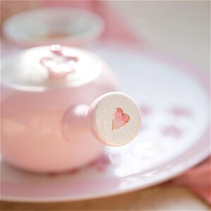 May include: A pink ceramic teapot with a heart-shaped detail on the handle. The teapot is sitting on a pink and white patterned plate.