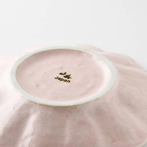 Pu&ograve; includere: Un piatto in ceramica rosa pallido con un bordo a forma di conchiglia. Il piatto ha un piccolo centro rotondo bianco con le parole "Japan" stampate in nero.