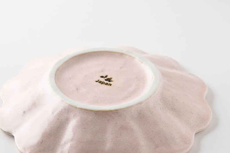 Pu&ograve; includere: Un piatto in ceramica rosa pallido con un bordo a forma di conchiglia. Il piatto ha un piccolo centro rotondo bianco con le parole "Japan" stampate in nero.