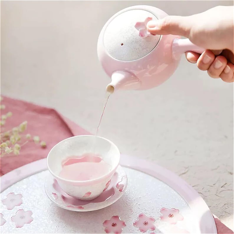 May include: A pink ceramic teapot with a floral design pours pink tea into a matching teacup and saucer. The teacup and saucer have a floral design and are on a white tray with a floral design.
