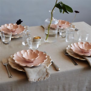 Pu&ograve; includere: Una tavola apparecchiata con una tovaglia color beige chiaro e piatti rosa a forma di fiore. I piatti sono impilati uno sopra l'altro e ci sono due forchette sul tavolo. C'&egrave; un vaso di vetro trasparente con uno stelo verde e foglie al centro del tavolo.
