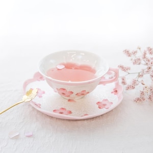 May include: A white teacup and saucer set with a pink floral design. The teacup is filled with pink liquid and sits on a matching saucer. A gold spoon rests on the saucer, and a sprig of pink flowers is in the background.