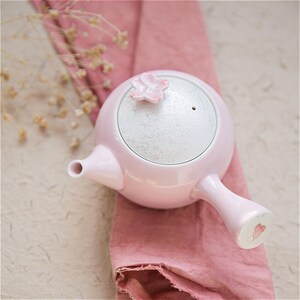 May include: A pink ceramic teapot with a white lid and a pink flower on top. The teapot has a small heart on the handle.