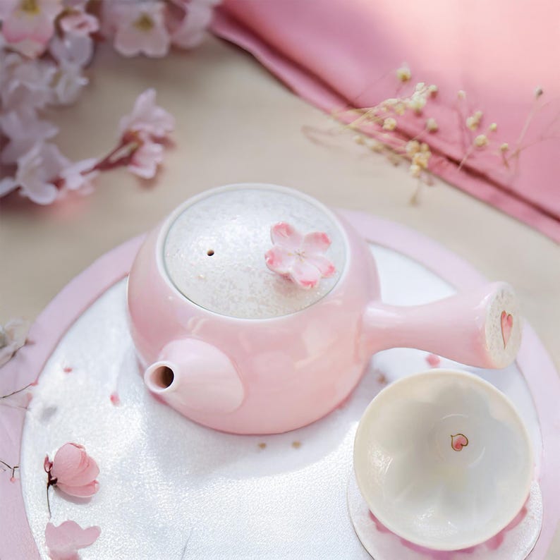 May include: A pink ceramic teapot with a white lid and a cherry blossom design. The teapot is sitting on a white tray with a cherry blossom pattern. A small pink teacup is next to the teapot.