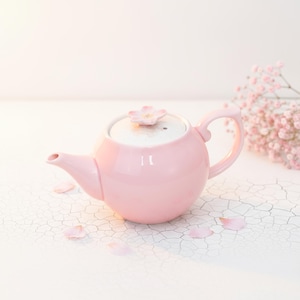 May include: A pink ceramic teapot with a flower-shaped lid handle. The teapot has a rounded body, a curved handle, and a spout. Scattered around the teapot are pink flower petals. The background is white.