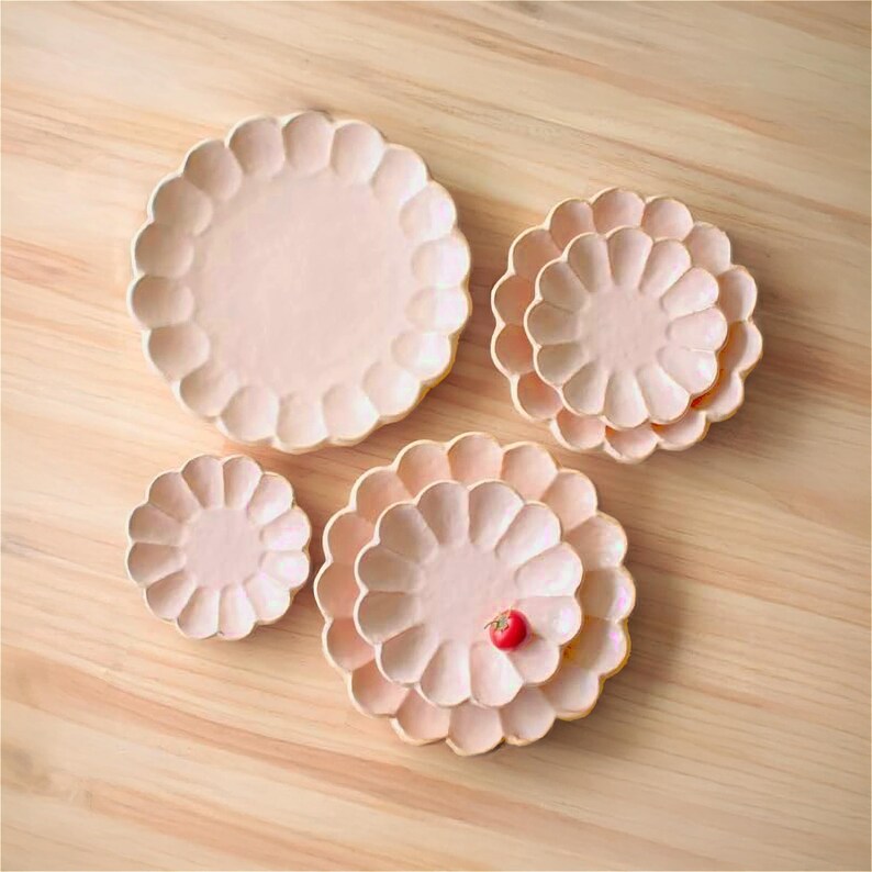 Pu&ograve; includere: Set di quattro piatti in ceramica rosa con bordi a festoni. I piatti sono disposti in ordine decrescente di dimensioni, con il piatto pi&ugrave; grande in alto e il pi&ugrave; piccolo in basso. Un piccolo pomodoro rosso &egrave; posto sul secondo piatto pi&ugrave; piccolo.