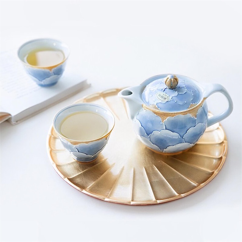 May include: A blue and gold ceramic teapot with a cloud design and a matching teacup and saucer. The teapot has a small knob on the lid. The teacup and saucer are on a gold tray.