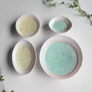 May include: Four ceramic plates and bowls in various sizes. The plates have a speckled, off-white rim with a pink accent. The interiors are light green and yellow. The largest plate is round, and the smallest is oval.