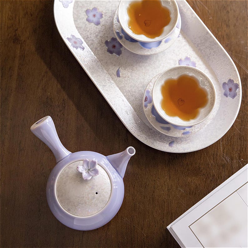 May include: A purple ceramic teapot with a floral design and a matching teacup and saucer. The teacup and saucer are on a white tray with a floral design.