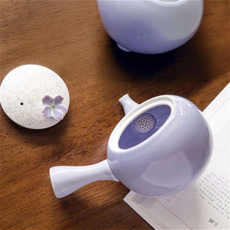 May include: A lavender ceramic teapot with a strainer and a matching lid. The teapot has a round body and a spout. The lid has a small flower design.