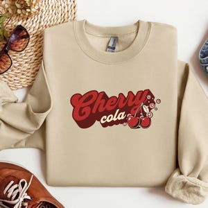 May include: A beige sweatshirt with a retro-style graphic design featuring the text "Cherry cola" and two red cherries with stems and leaves.
