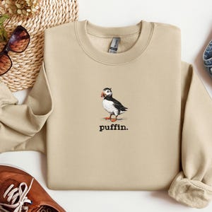 May include: A beige sweatshirt featuring a graphic of a puffin with the word "puffin." printed beneath it.