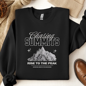 Könnte beinhalten: Schwarzer Sweatshirt mit einem weißen Grafikdesign einer Bergkette und dem Text "Chasing Summits Rise to the Peak Adventure Awaits in the Mountains".