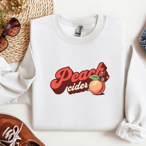 May include: White crew neck sweatshirt featuring the text "Peach Cider" in a retro style, with a peach graphic. The image includes sunglasses, denim jeans, and brown shoes.