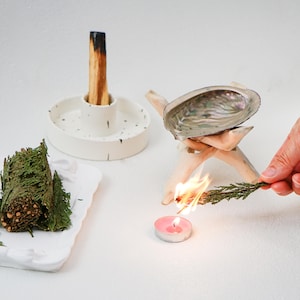 May include: A white ceramic holder with a wooden stick incense, a white ceramic dish with a green bundle of sage, a white ceramic dish with a pink candle, and a seashell on a wooden stand with a lit sage bundle.