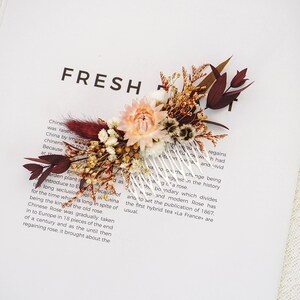Puede incluir: Un peine floral para el cabello con un grupo de flores secas en tonos melocotón, burdeos y marrón. El peine es de plástico transparente y está sobre un fondo blanco con la palabra "FRESH" impresa.