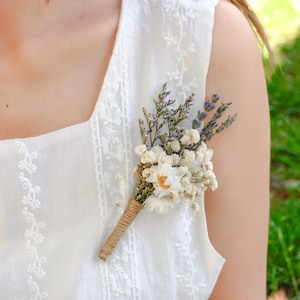 May include: A white sleeveless dress with delicate embroidery and a small bouquet of dried flowers pinned to the front.
