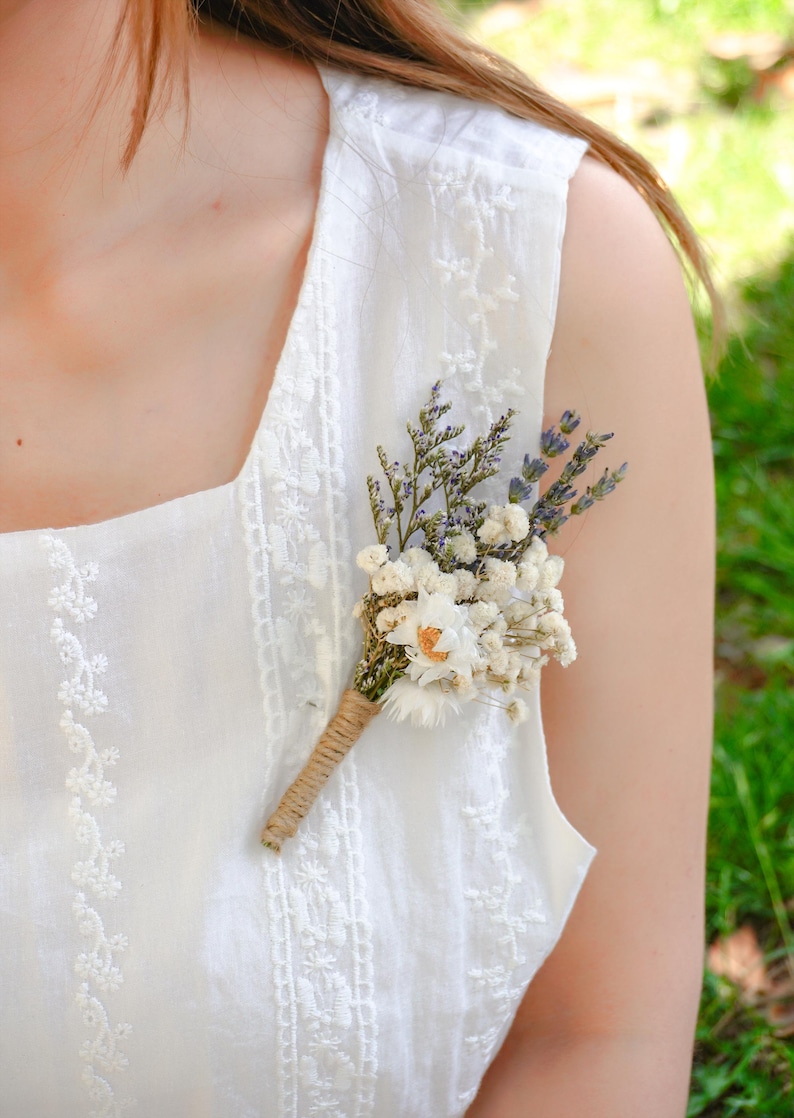 May include: A white sleeveless dress with delicate embroidery and a small bouquet of dried flowers pinned to the front.