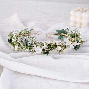 May include: A floral crown with white flowers, green leaves, and sprigs of greenery. The crown has a thin, gold-coloured wire base and is tied with a white ribbon. The crown is suitable for special occasions.