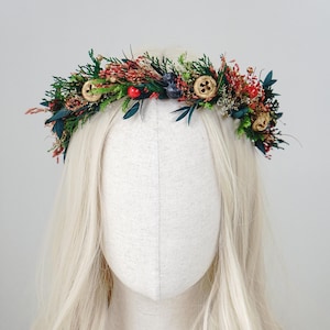 Puede incluir: Una corona floral hecha a mano con una mezcla de flores secas y vegetación. La corona incluye bayas rojas, hojas verdes oscuras y pequeños detalles de madera. Se muestra en una cabeza de maniquí con pelo largo y rubio.