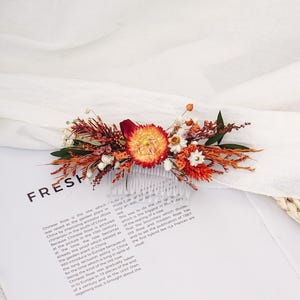 Puede incluir: Un peine floral para el cabello con flores secas en tonos naranja, rojo y blanco. El peine es de plástico transparente y es ideal para bodas u ocasiones especiales.