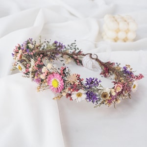 May include: A circular floral crown constructed from a variety of dried flowers in shades of pink, purple, white, and yellow. The arrangement features small daisies, strawflowers, and sprigs of greenery attached to a thin, flexible brown vine base.